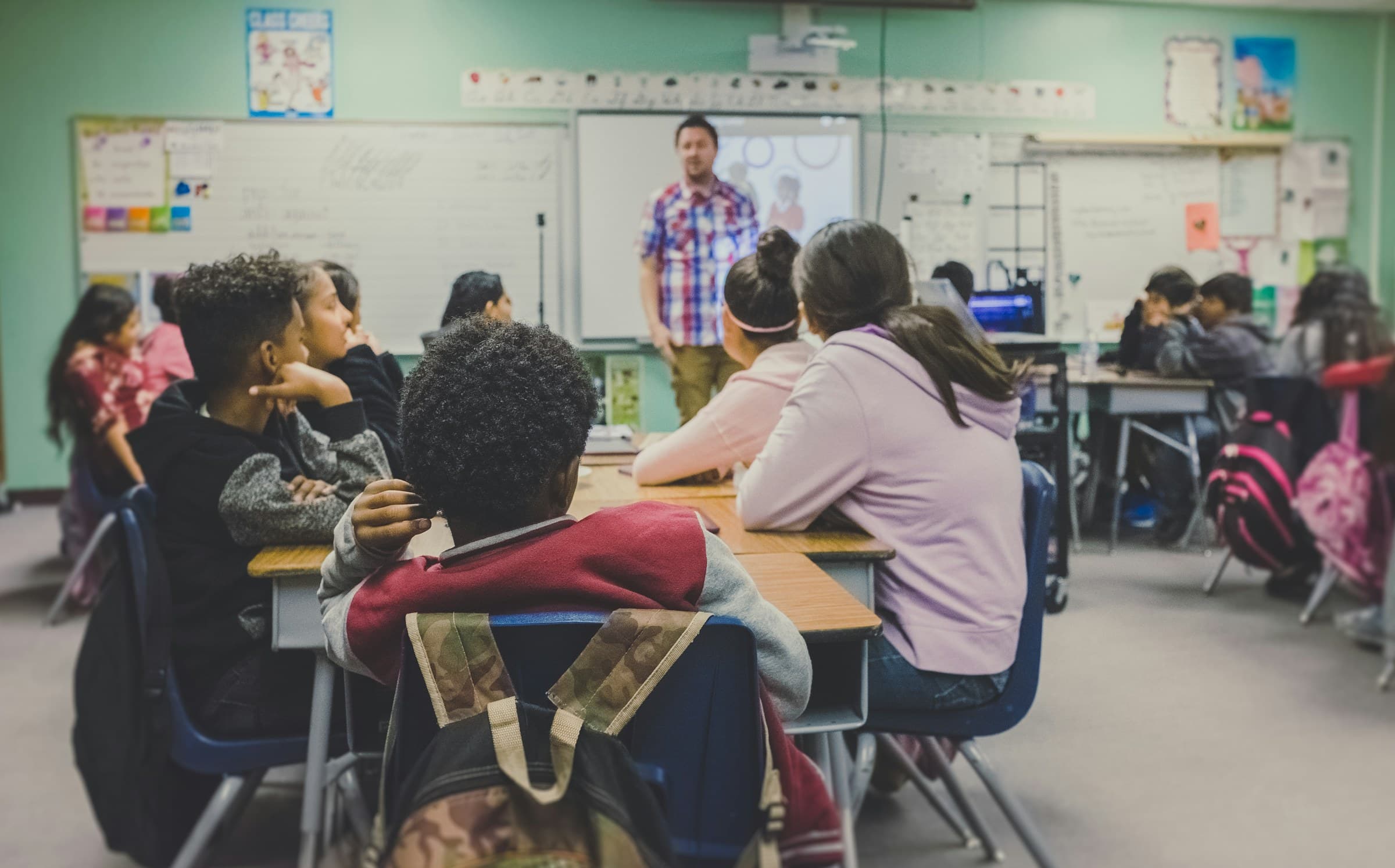 A teacher leads a class of middle school students seated at group tables in a school classroom.
